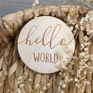 May include: A round wooden sign with the words "hello world" engraved in a cursive font. The sign is sitting on a woven basket with dried white flowers.