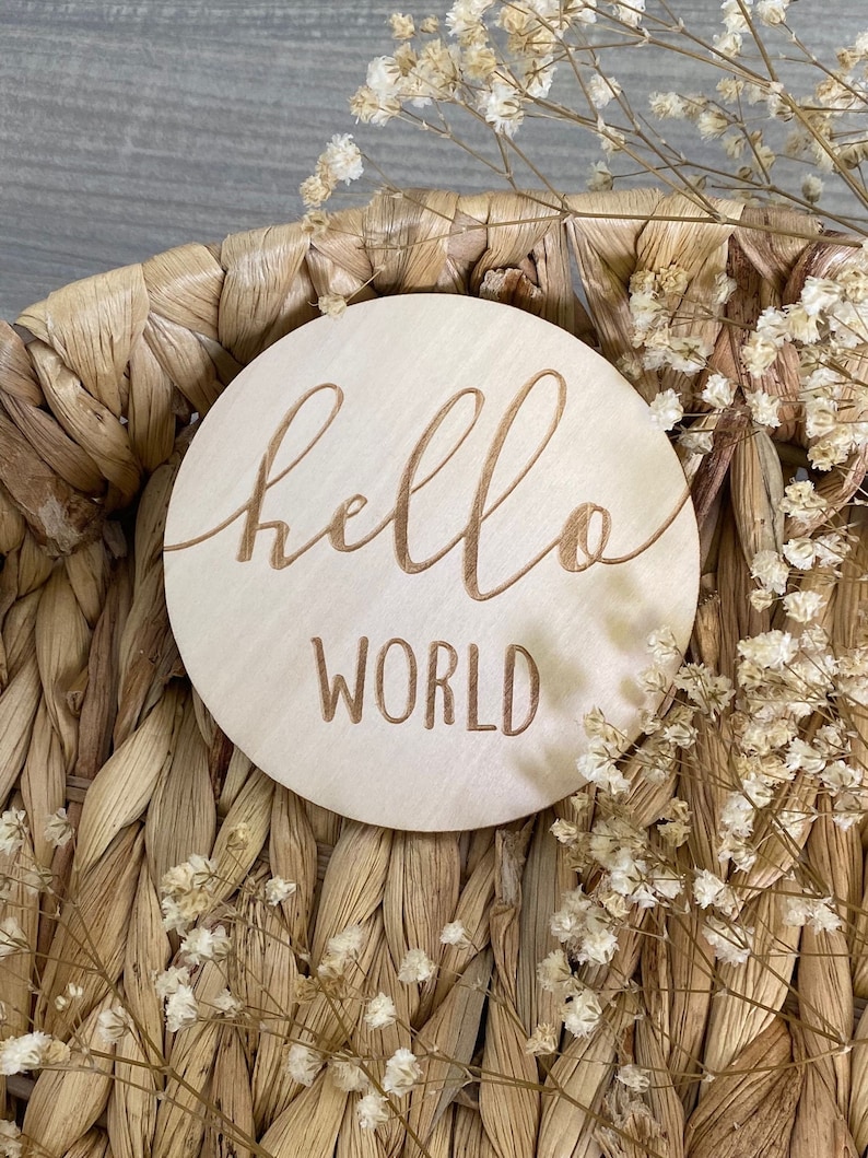 May include: A round wooden sign with the words "hello world" engraved in a cursive font. The sign is sitting on a woven basket with dried white flowers.