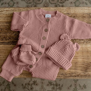 May include: A dusty rose knitted baby set consisting of a long-sleeved button-down romper, a pair of matching booties, and a beanie with small bear ears. The romper features a ribbed texture, a round neckline, and a label at the inner collar.