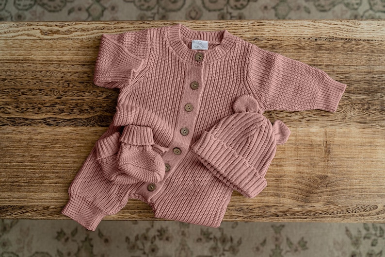 May include: A dusty rose knitted baby set consisting of a long-sleeved button-down romper, a pair of matching booties, and a beanie with small bear ears. The romper features a ribbed texture, a round neckline, and a label at the inner collar.
