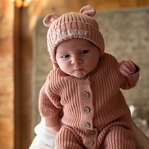 May include: A dusty rose knitted beanie with small bear ears and the name Eleanor embroidered in white script. The beanie is paired with a matching long-sleeve knitted romper featuring a vertical ribbed texture and five round wooden buttons.