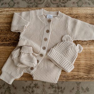 May include: A beige knitted baby romper featuring a ribbed texture, long sleeves, and a front button closure. The set includes a matching pair of knitted booties and a beanie hat with two small rounded ears on top, all crafted in a neutral tone.
