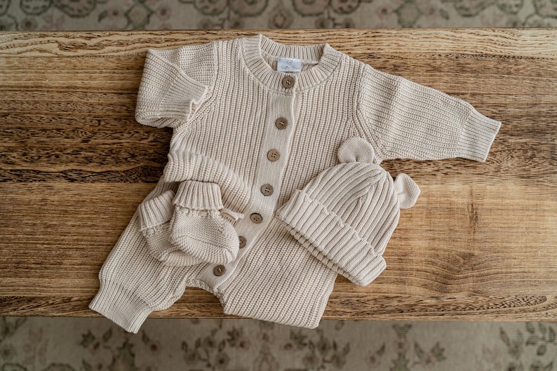 May include: A beige knitted baby romper featuring a ribbed texture, long sleeves, and a front button closure. The set includes a matching pair of knitted booties and a beanie hat with two small rounded ears on top, all crafted in a neutral tone.
