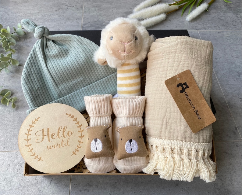 May include: A gift box filled with baby items, including a light blue knitted hat, a wooden sign that says "Hello world", a pair of cream-colored socks with brown bear faces, a cream-colored muslin blanket with fringe, and a plush lamb rattle with yellow and white stripes.