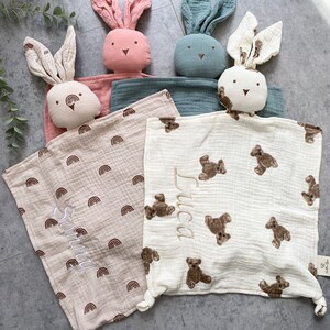 May include: Three personalized baby blankets with bunny-shaped security toys. The blankets are made of soft, white fabric with different patterns. One blanket has a rainbow pattern, another has a bear pattern, and the third has a bear pattern with the name "Luca" embroidered on it.