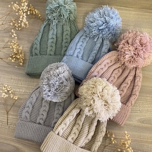 May include: Five knitted hats with pom-poms in various colors, including gray, blue, green, beige, and pink. The hats are made with a cable knit pattern and have a fluffy pom-pom on top.
