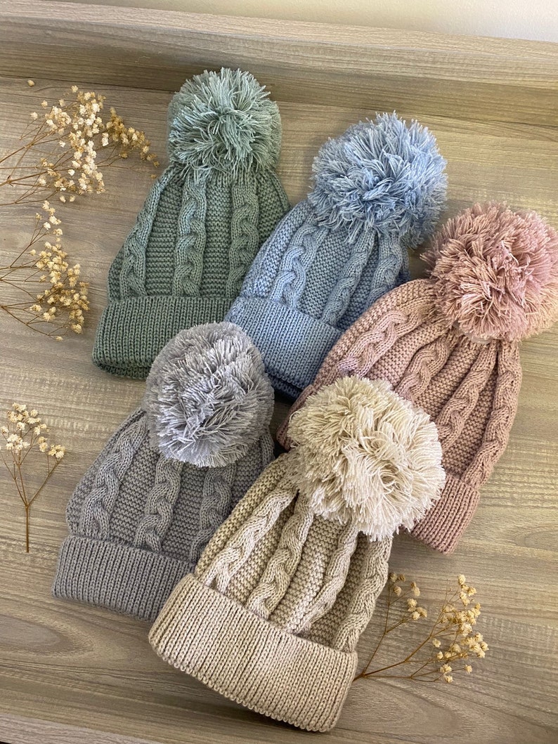 May include: Five knitted hats with pom-poms in various colors, including gray, blue, green, beige, and pink. The hats are made with a cable knit pattern and have a fluffy pom-pom on top.