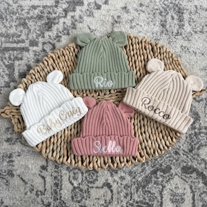 May include: Four ribbed knit beanies with bear ears, each featuring embroidered names. The hats are white with 'Baby Craig', sage green with 'Rio', dusty rose with 'Stella', and beige with 'Rocco'. Each beanie has a folded cuff design.