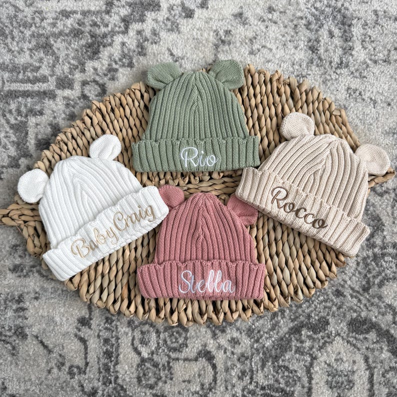 May include: Four ribbed knit beanies with bear ears, each featuring embroidered names. The hats are white with 'Baby Craig', sage green with 'Rio', dusty rose with 'Stella', and beige with 'Rocco'. Each beanie has a folded cuff design.