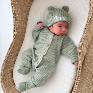 May include: A sage green knitted long-sleeve romper with a front button closure and matching booties. The outfit includes a ribbed beanie with small bear ears and the name Isaac embroidered in white script on the front of the hat.