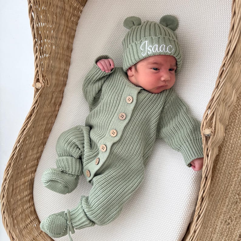 May include: A sage green knitted long-sleeve romper with a front button closure and matching booties. The outfit includes a ribbed beanie with small bear ears and the name Isaac embroidered in white script on the front of the hat.