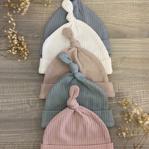 May include: Five knitted baby hats in different colors: light blue, white, beige, dark blue, and pink. The hats are stacked on top of each other, with the pink hat on the bottom.