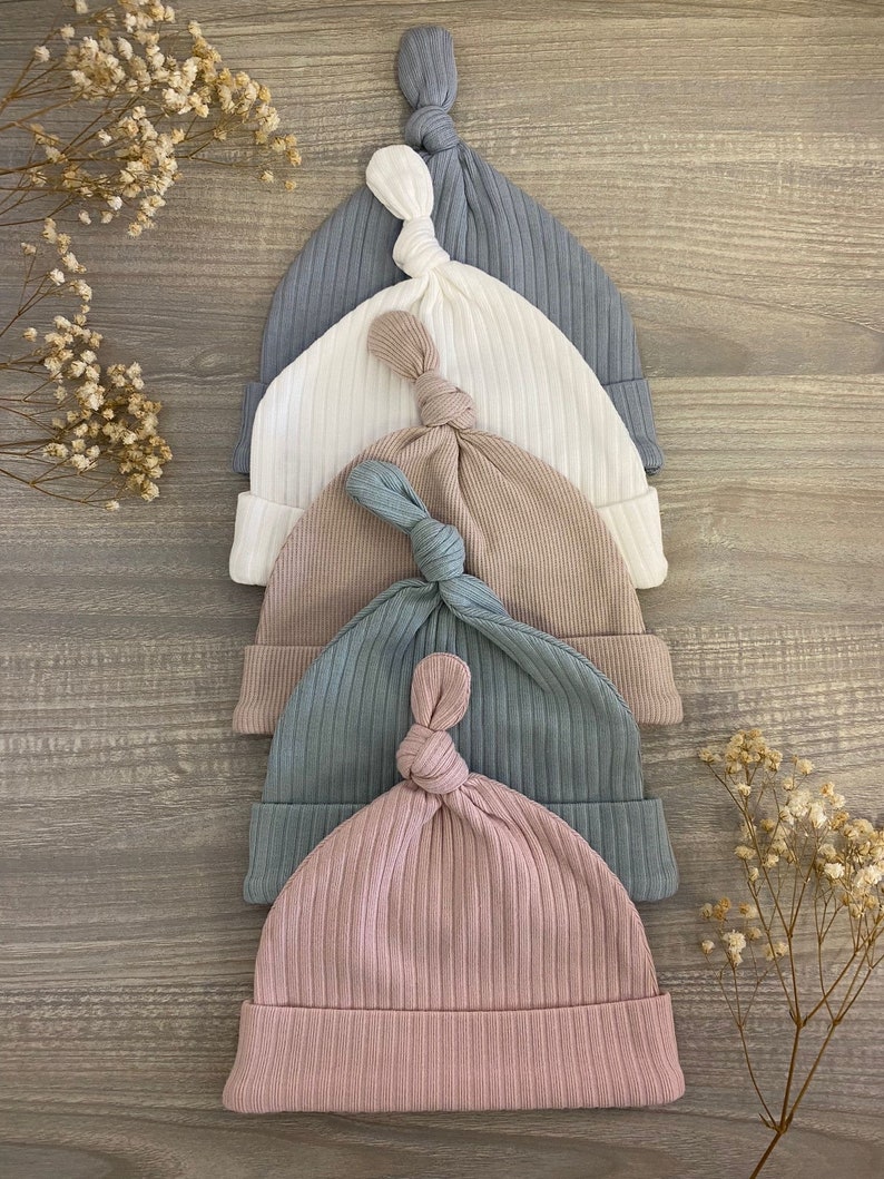 May include: Five knitted baby hats in different colors: light blue, white, beige, dark blue, and pink. The hats are stacked on top of each other, with the pink hat on the bottom.