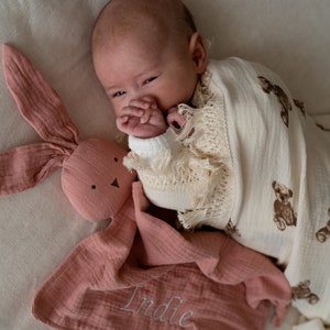 May include: A pink muslin baby blanket with the name "Indie" embroidered on it. A baby is wrapped in a white muslin blanket with brown bear prints and is holding a pink bunny lovey.