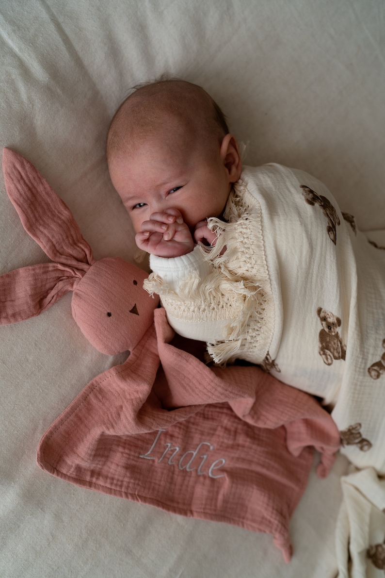 May include: A pink muslin baby blanket with the name "Indie" embroidered on it. A baby is wrapped in a white muslin blanket with brown bear prints and is holding a pink bunny lovey.