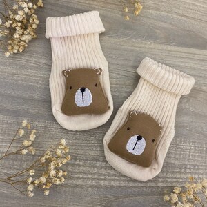 May include: A pair of cream-colored baby socks with brown bear faces on the toes. The socks are made of soft, knitted fabric and are perfect for keeping little feet warm.