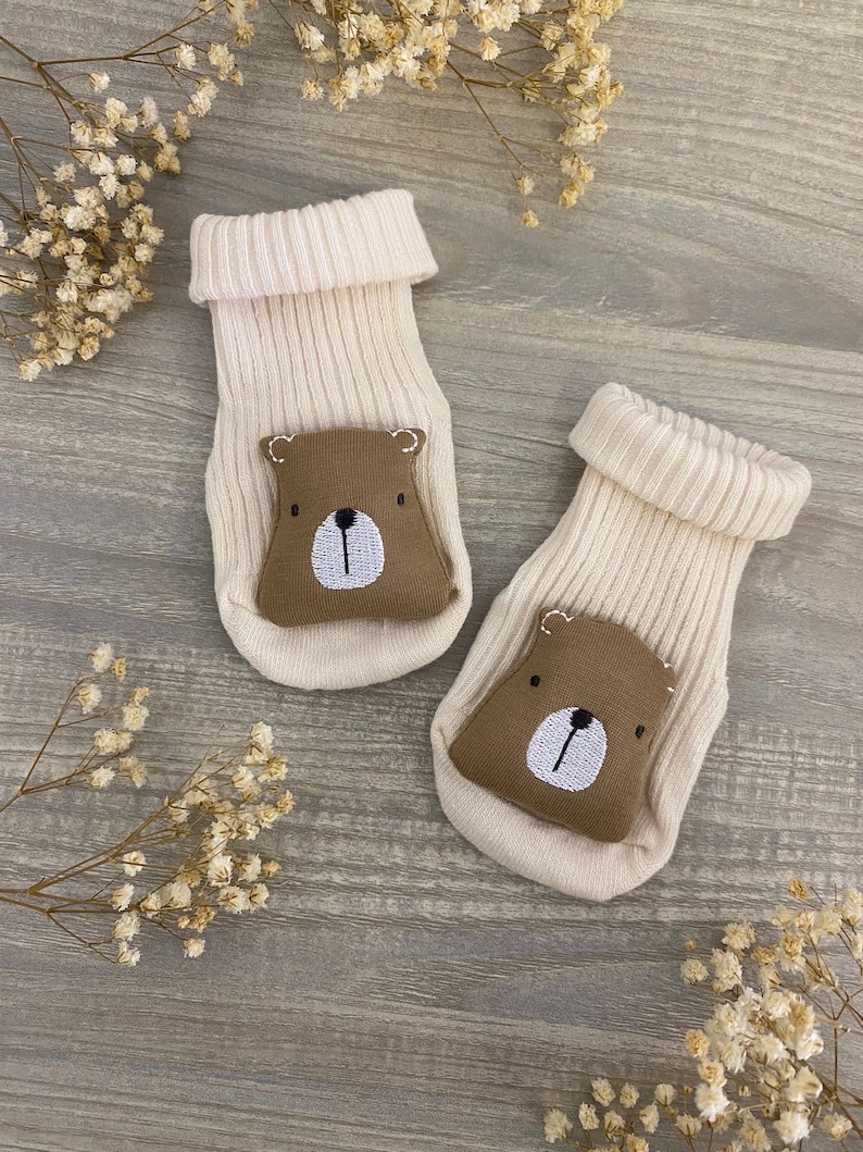 May include: A pair of cream-colored baby socks with brown bear faces on the toes. The socks are made of soft, knitted fabric and are perfect for keeping little feet warm.