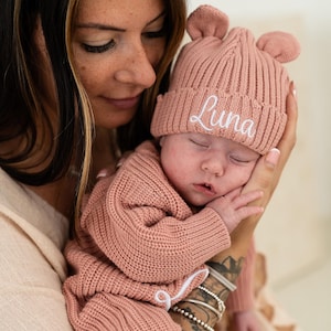 May include: A dusty rose knitted baby beanie featuring two small rounded ears on top and the name Luna embroidered in white cursive script on the folded cuff. The hat is made of a thick ribbed knit fabric and matches a long-sleeved knitted outfit.