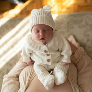 May include: A cream-colored ribbed knit outfit consisting of a long-sleeved button-down cardigan, matching pants, and a beanie with small bear ears. The cardigan features four brown buttons down the front, and the set is made of a soft, textured yarn.