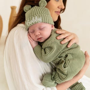 May include: A sage green knitted baby beanie with small bear ears and the name Bobby embroidered in white script on the folded cuff, paired with a matching sage green long-sleeved knitted baby romper with integrated footies and ribbed texture.