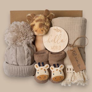 May include: A baby gift set in a cardboard box. The set includes a giraffe plush toy, a knitted hat with a pom-pom, a blanket, a wooden sign that says "hello world", and baby socks with giraffe faces.
