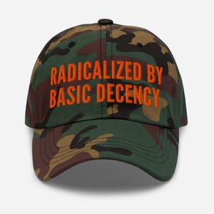 Op de afbeelding: Camouflage baseballpet met de tekst "RADICALIZED BY BASIC DECENCY" in oranje blokletters. De pet heeft een gebogen klep en een zwarte knoop. De hoed is een mix van groen, bruin en zwart.