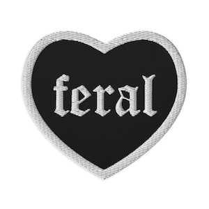May include: A heart-shaped embroidered patch with a black background and a white border. The word "feral" is stitched in the center in a white, gothic-style font. The patch features a clean, stitched edge that defines the heart shape clearly.