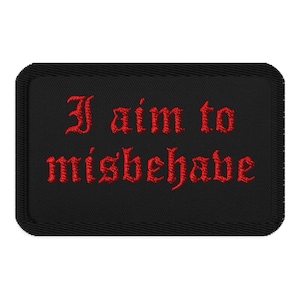 Puede incluir: Un parche rectangular negro bordado con un borde negro grueso. El texto "I aim to misbehave" está cosido en una fuente gótica roja y llamativa en el centro del parche, creando un diseño de alto contraste sobre el fondo de tela oscura.