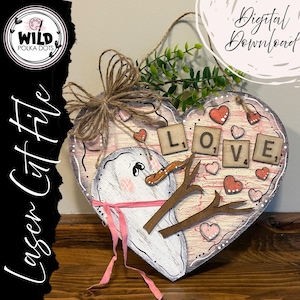 以下が含まれることがあります： 雪だるまのデザインと「LOVE」の文字がスクラブルタイルで書かれた、ハート型の木製サイン。ピンクと白のアクセント、小さなハート、ジュート紐のハンガーでペイントされています。「Laser Cut File」と「Digital Download」の文字も表示されています。