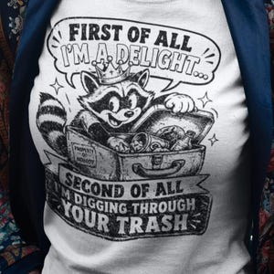 May include: A white t-shirt featuring a black graphic of a raccoon wearing a crown, sitting in a trash can. The text reads: FIRST OF ALL I'M A DELIGHT... SECOND OF ALL I'M DIGGING THROUGH YOUR TRASH. The shirt is worn under a dark blue floral jacket.