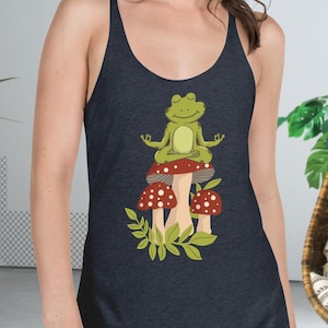 May include: Dark navy blue tank top featuring a cartoon frog meditating on top of mushrooms. The frog is green, and the mushrooms are red with white spots. The design includes green leaves at the base of the mushrooms.