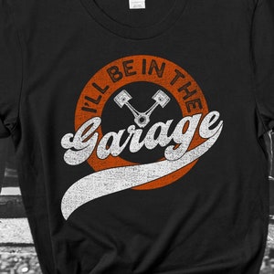 K&ouml;nnte beinhalten: Schwarzes T-Shirt mit dem Text "I'll Be In The Garage" in einem Retro-Design. Das Design umfasst einen orangefarbenen Kreis mit dem Text und zwei gekreuzten Kolben. Das Shirt ist von der Marke Gildan.
