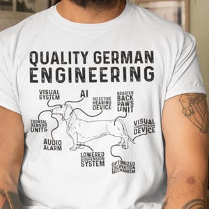May include: A white short-sleeved t-shirt featuring a black graphic of a dachshund with technical labels. The text reads Quality German Engineering, AI, Visual System, Selective Hearing Device, Booster Paws Unit, Frontal Sensor Units, Audio Alarm, Lowered Suspension System, and Optimized Excavation Mechanism.