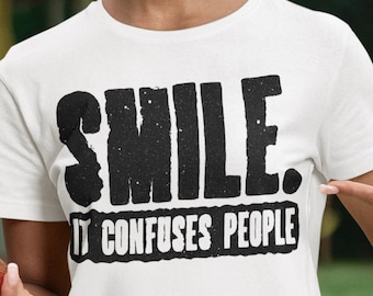 May include: A white short-sleeved t-shirt featuring the black text "SMILE. IT CONFUSES PEOPLE" printed in a bold, distressed font across the chest. The shirt has a classic crew neckline and a straight hem, made of a plain white fabric.