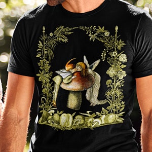 K&ouml;nnte beinhalten: Schwarzes T-Shirt mit einer skurrilen Illustration eines Feenpaares auf einem Pilz, umrahmt von einem floralen Bogen. Das Design verwendet Gr&uuml;n-, Braun- und Beiget&ouml;ne. Das Paar h&auml;lt ein Buch. Das T-Shirt wird von einer Person getragen.