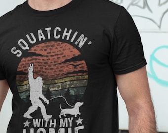 May include: A black short-sleeved t-shirt featuring a graphic of a Bigfoot walking a dachshund. The design includes a distressed sunset background and the text "SQUATCHIN' WITH MY HOMIE" in white lettering, with stars flanking the word "WITH".