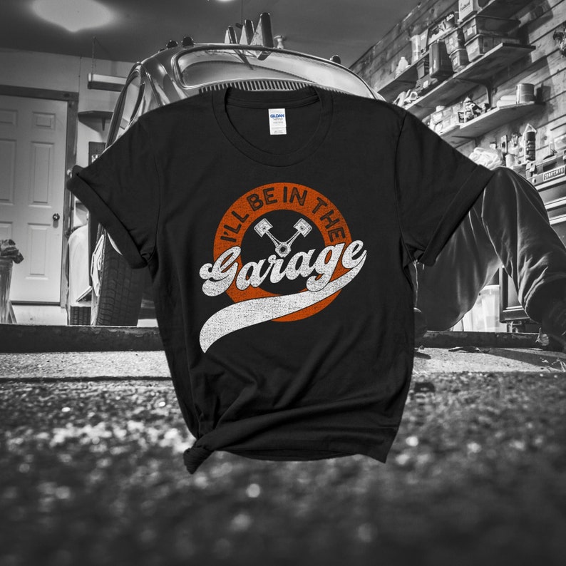 K&ouml;nnte beinhalten: Schwarzes T-Shirt mit dem Text "I'll Be In The Garage" in einem Retro-Design. Das Design umfasst einen orangefarbenen Kreis mit dem Text und zwei gekreuzten Kolben. Das Shirt ist von der Marke Gildan.