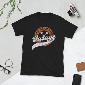 K&ouml;nnte beinhalten: Schwarzes Kurzarm-T-Shirt mit einem verblichenen orangefarbenen und wei&szlig;en Aufdruck. Der Aufdruck lautet "I'll Be In The Garage" mit einer Vintage-Schriftart und zwei Kolben-Illustrationen. Eine Kamera, eine Brieftasche und ein Telefon sind sichtbar.