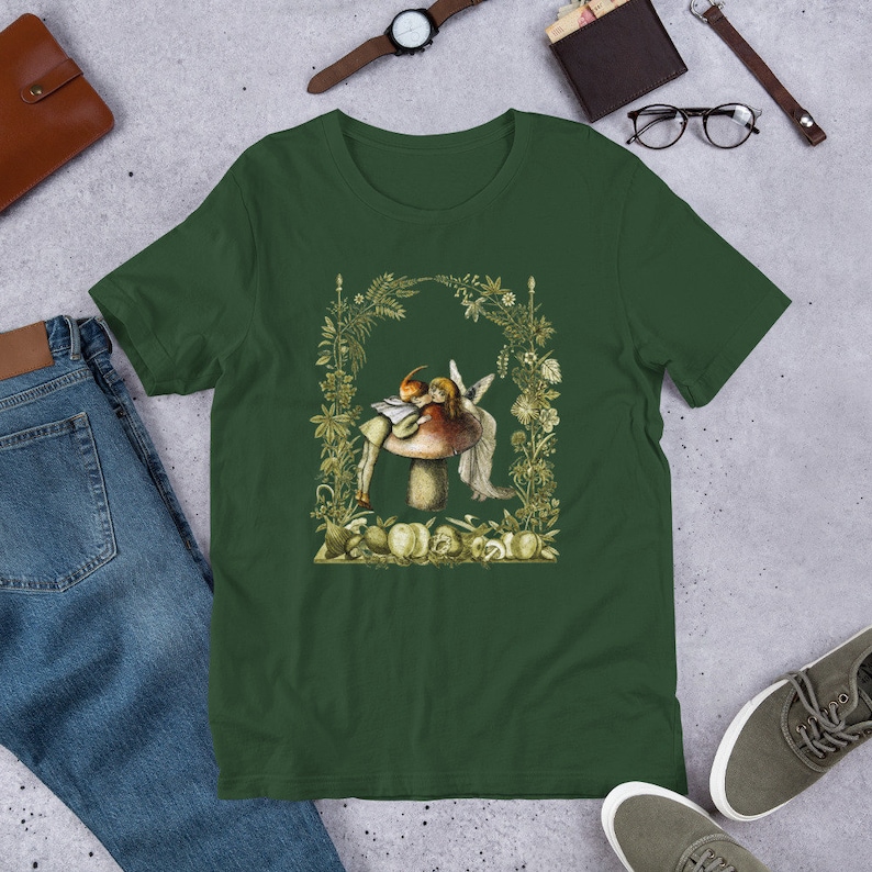 May include: Dark green t-shirt featuring a whimsical illustration of a fairy and a mushroom. The design is framed by a floral arch and includes a variety of fruits and foliage. The shirt is made of a soft material.