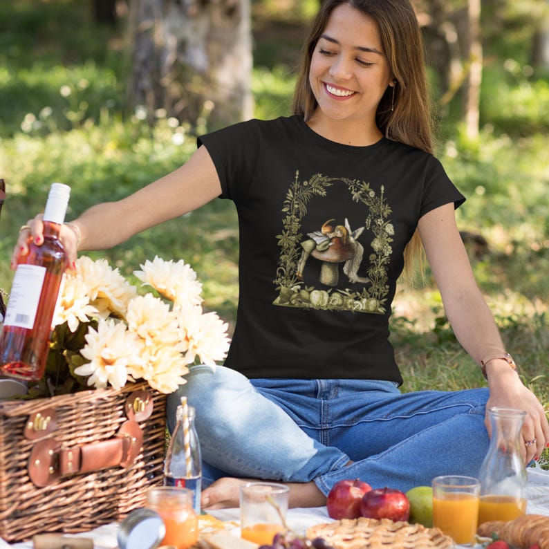 K&ouml;nnte beinhalten: Schwarzes T-Shirt mit einer Illustration einer Fee und eines Pilzes, umrahmt von einem floralen Bogen. Die Person sitzt auf dem Gras mit einem Picknickkorb, Blumen und einer Weinflasche. Picknick-Szene.