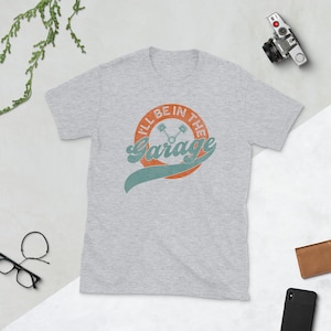 K&ouml;nnte beinhalten: Hellgraues T-Shirt mit dem Text "I'll Be In The Garage" in einem Retro-Design. Das Design zeigt einen orangefarbenen Kreis mit Text, ein t&uuml;rkisfarbenes Banner und gekreuzte Kolben. Eine Vintage-Kamera und eine Brieftasche sind im Hintergrund.