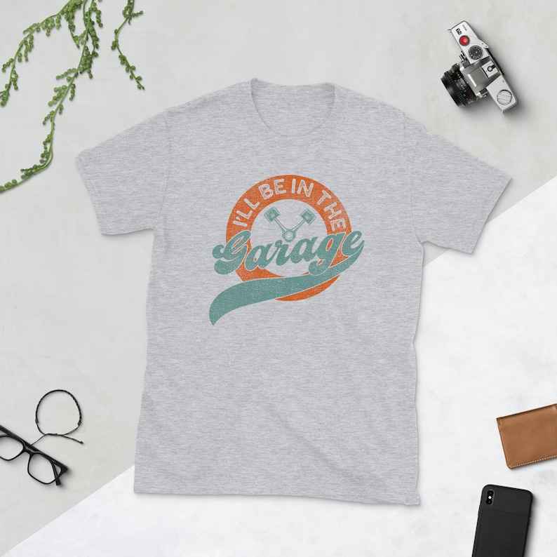 K&ouml;nnte beinhalten: Hellgraues T-Shirt mit dem Text "I'll Be In The Garage" in einem Retro-Design. Das Design zeigt einen orangefarbenen Kreis mit Text, ein t&uuml;rkisfarbenes Banner und gekreuzte Kolben. Eine Vintage-Kamera und eine Brieftasche sind im Hintergrund.