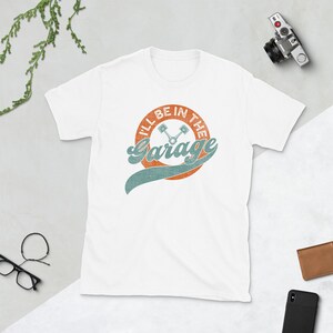 K&ouml;nnte beinhalten: Wei&szlig;es T-Shirt mit Retro-Grafik. Das Design zeigt den Text "I'LL BE IN THE Garage" im Vintage-Stil, mit einem orangefarbenen Kreis und t&uuml;rkisfarbenen Akzenten. Zwei gekreuzte Kolben befinden sich in der Mitte.