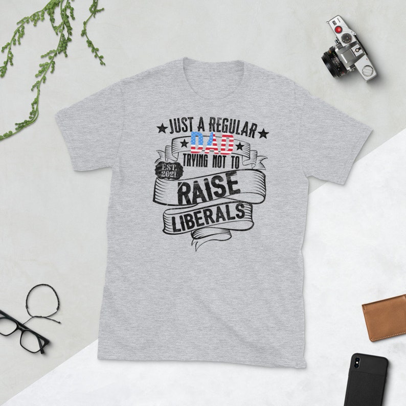 Op de afbeelding: Lichtgrijs t-shirt met de tekst "JUST A REGULAR DAD TRYING NOT TO RAISE LIBERALS" in zwart en rood. Het ontwerp bevat een Amerikaanse vlag en de tekst "EST. 2021".