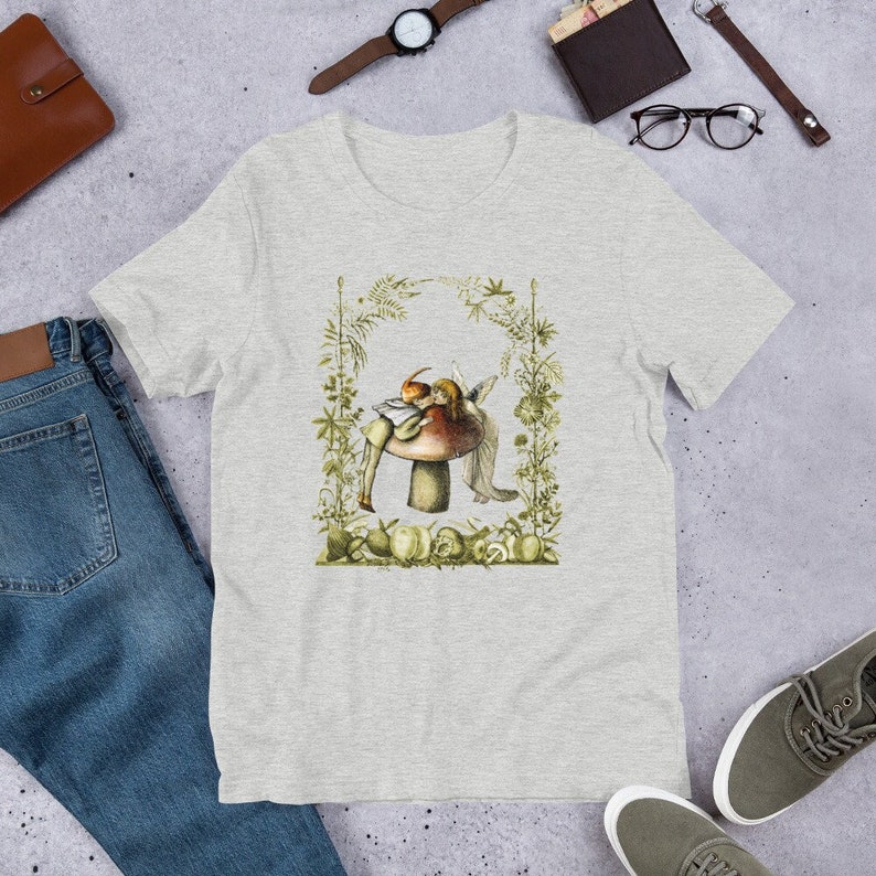 K&ouml;nnte beinhalten: Hellgraues T-Shirt mit einer Fantasy-Illustration von zwei Feen auf einem Pilz, umrahmt von gr&uuml;nen Bl&auml;ttern und Pflanzen. Das Design beinhaltet verschiedene Fr&uuml;chte und Gem&uuml;se an der Basis. Das T-Shirt ist aus weichem Material.