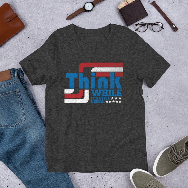 May include: A dark heather grey t-shirt with a distressed American flag design and the text "Think While It's Still Legal" in white.