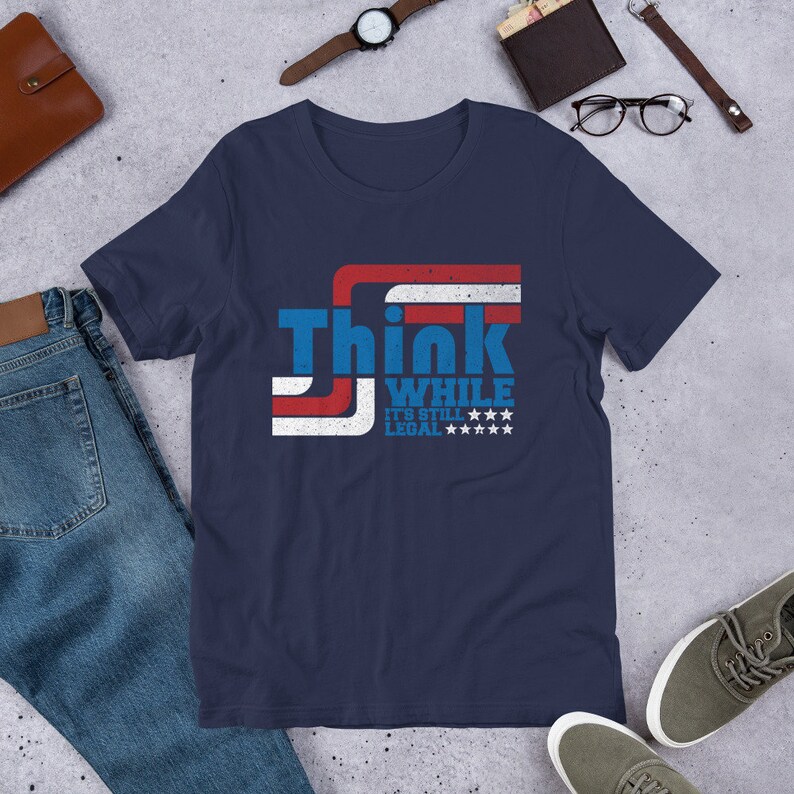 May include: Navy blue t-shirt with a red, white, and blue American flag design. The shirt has the text "Think While It's Still Legal" printed on it.