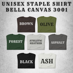 K&ouml;nnte beinhalten: Auswahl an Unisex-Basic-Shirts in verschiedenen Farben, darunter Braun, Olive, Waldgr&uuml;n, Athletic Heather, Asphalt, Schwarz und Asch. Jedes Shirt ist gefaltet und mit seinem Farbnamen in fetter wei&szlig;er Schrift versehen. Der Text "UNISEX STAPLE SHIRT BELLA CANVAS 3001" steht oben.