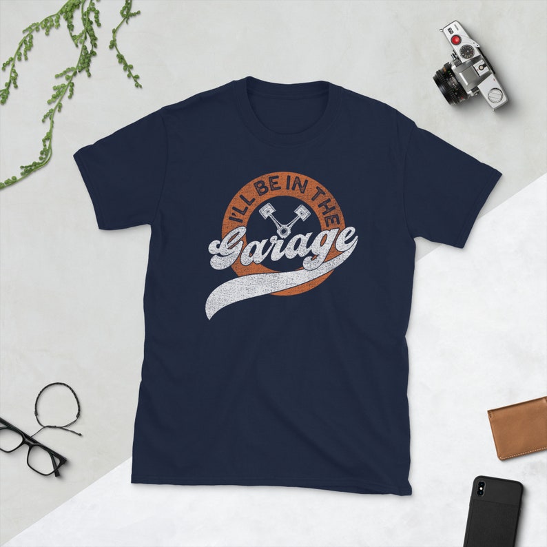 K&ouml;nnte beinhalten: Marineblaues T-Shirt mit dem Text "I'll Be In The Garage" in einem Retro-Design. Das Design zeigt einen orangefarbenen Kreis mit dem Text und zwei Kolben-Grafiken. Das Shirt hat einen Rundhalsausschnitt und kurze &Auml;rmel.