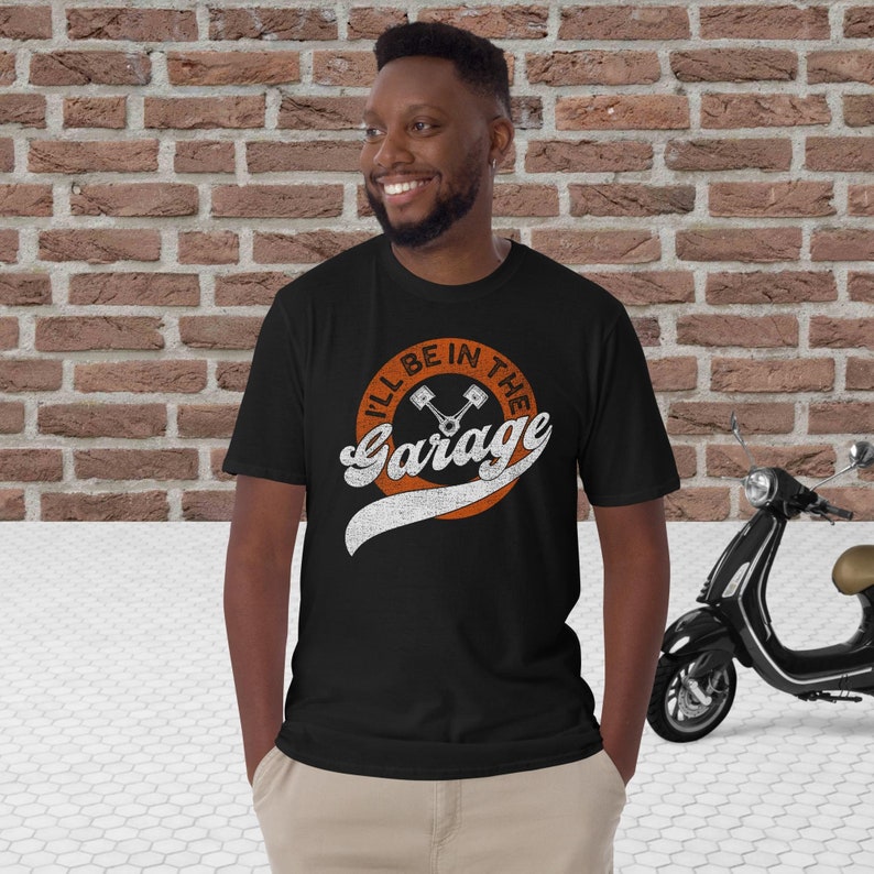K&ouml;nnte beinhalten: Schwarzes T-Shirt mit dem Text "I'll Be In The Garage" in einem verwitterten orange-wei&szlig;en Design. Die Grafik enth&auml;lt gekreuzte Kolben in einem orangefarbenen Kreis. Im Hintergrund ist ein Roller im Vintage-Stil zu sehen.